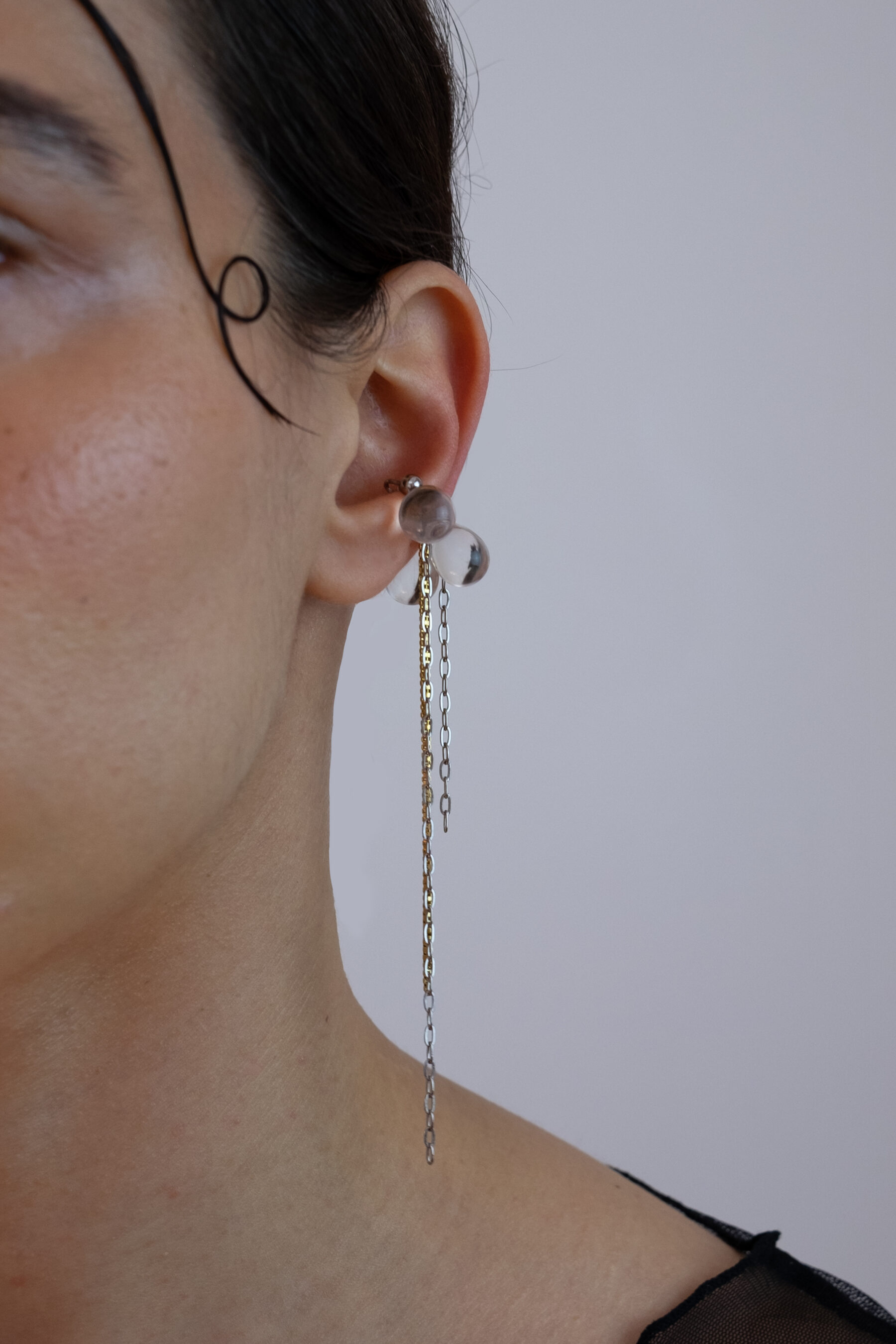 Earcuff en acier inoxydable. Il s’agit d’un faux piercing pour oreille non perçée qui se positionne au niveau du conch ou du lobe suivant la largeur de l’oreille. Il est composé d’un anneau argenté, de perles en verre en forme de goutte transparentes et de chaines dorées et argentées pendantes.